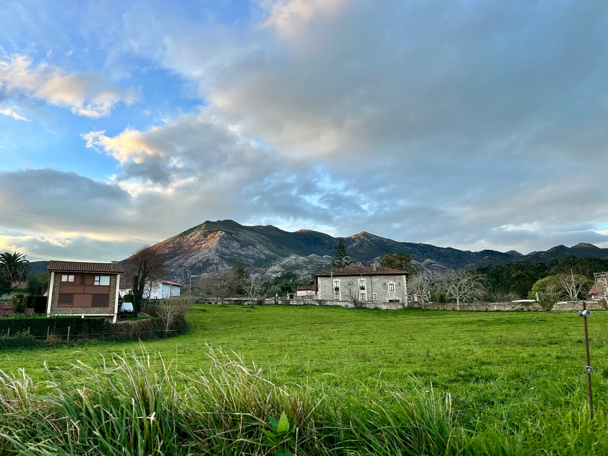 Prado verde en primer plano con caseríos de piedra y montañas al fondo bajo un cielo azul con nubes al amanecer en Caravia, Asturias