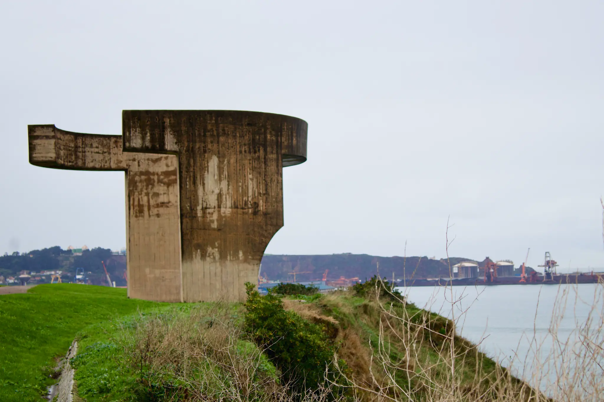 Escultura de hormigón en forma de proa de barco sobre acantilado con vistas al puerto industrial de Gijón y la costa asturiana