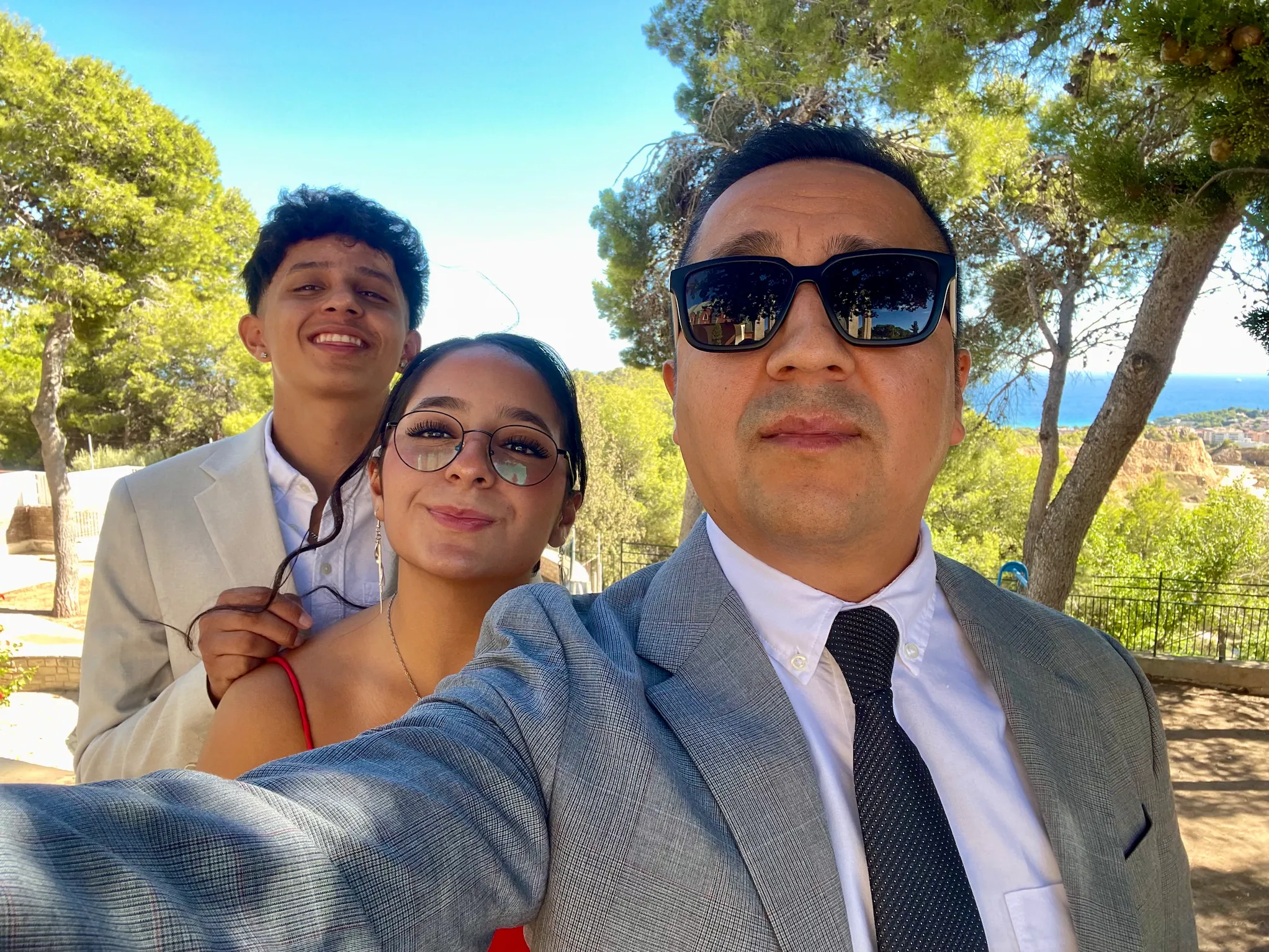 Selfie de tres personas vestidas para una boda, sonrientes bajo los árboles antes de la ceremonia.