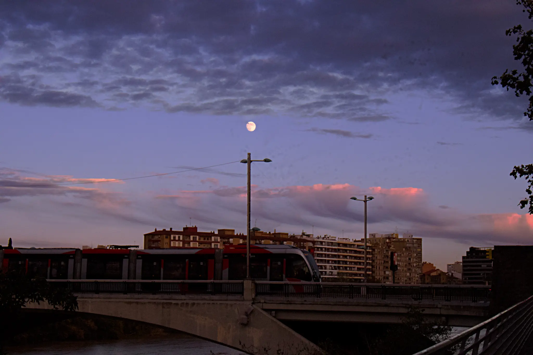 Luna llena asomando sobre el puente de Zaragoza durante un atardecer con nubes rosadas