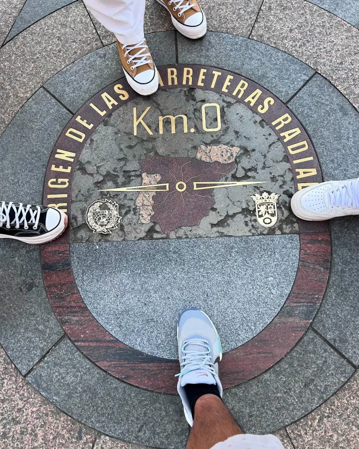 Familia de cuatro posando con sus zapatillas sobre la placa del Km 0 en la Puerta del Sol de Madrid, punto de origen de las carreteras radiales de España.