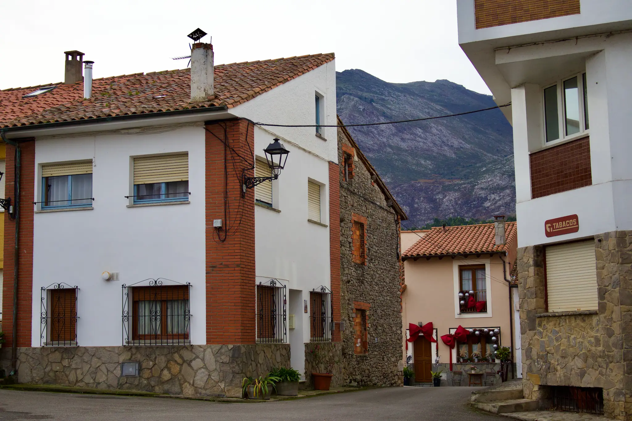 Calle de Duesos con casas rústicas de piedra, ladrillo y teja, ventanas con rejas de forja y montaña al fondo bajo cielo nublado en Asturias