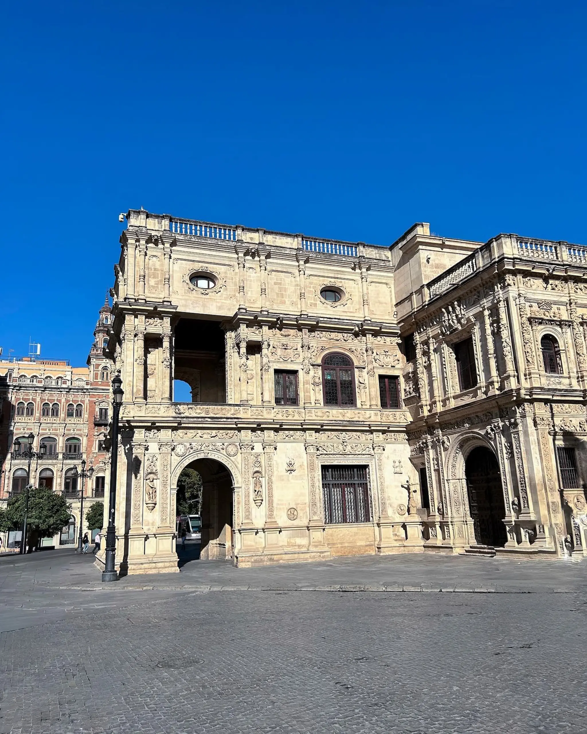 Fachada plateresca del Ayuntamiento de Sevilla iluminada por el sol en la Plaza de San Francisco, con cielo azul despejado y la plaza prácticamente vacía.